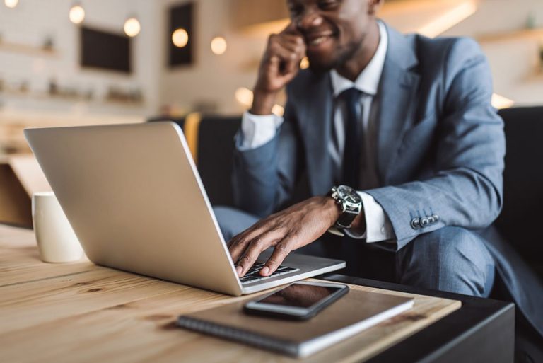 Un homme d'affaires souriant en costume travaillant sur des stratégies de référencement sur un ordinateur portable installé sur un bureau en bois, avec un smartphone et un ordinateur portable à proximité.