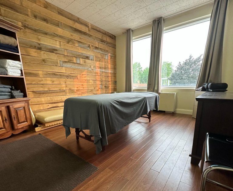 Un accueil dans une salle de massothérapie avec une table de massage recouverte d'un drap gris, des boiseries murales rustiques, une fenêtre et des étagères bien organisées.